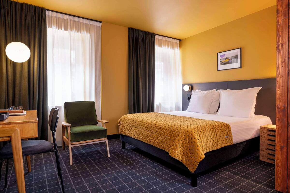 A hotel room with a bed, a chair, a desk, and a lamp.