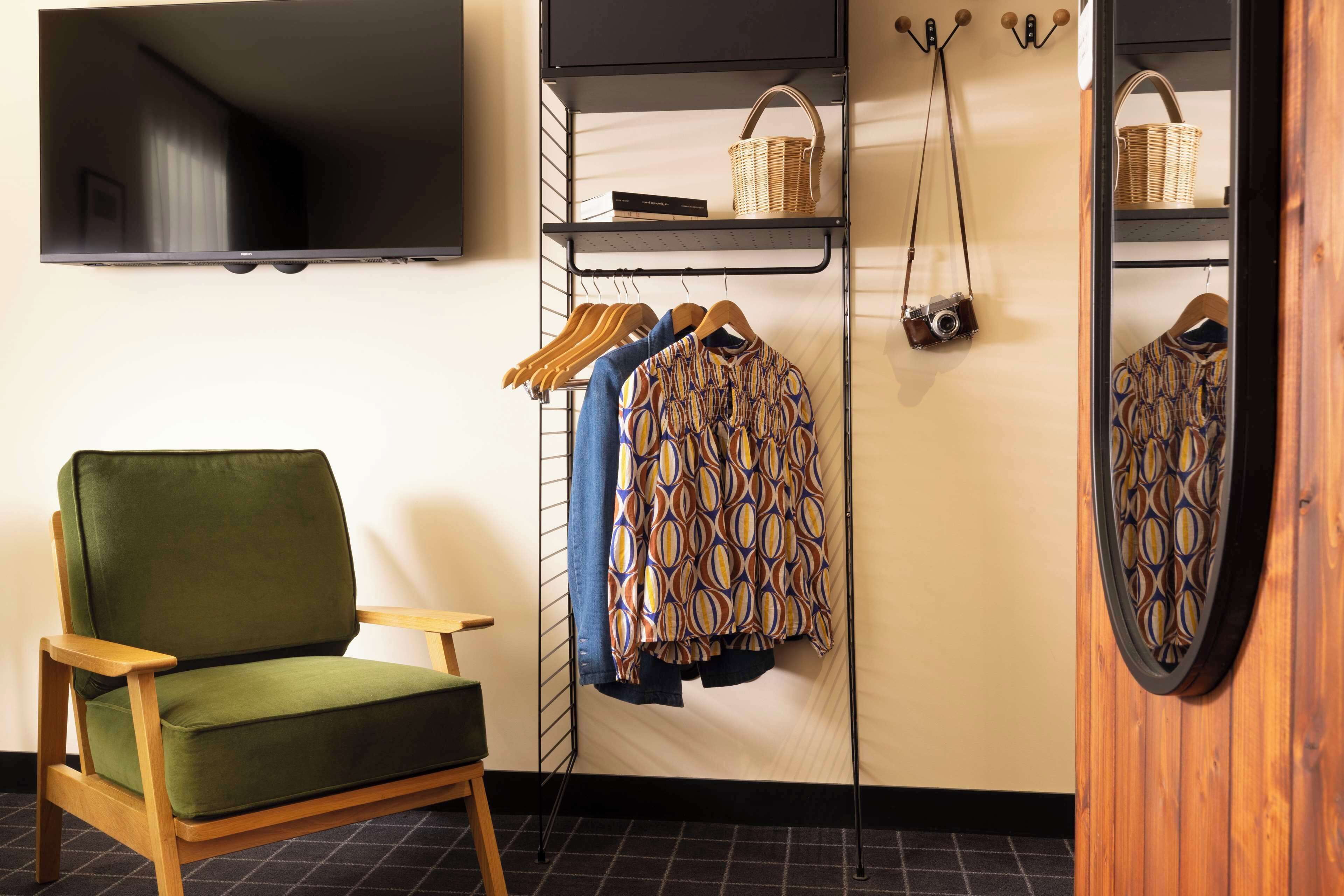 A room with a green armchair, a wardrobe with hanging clothes, a shelf with a basket, and a camera.