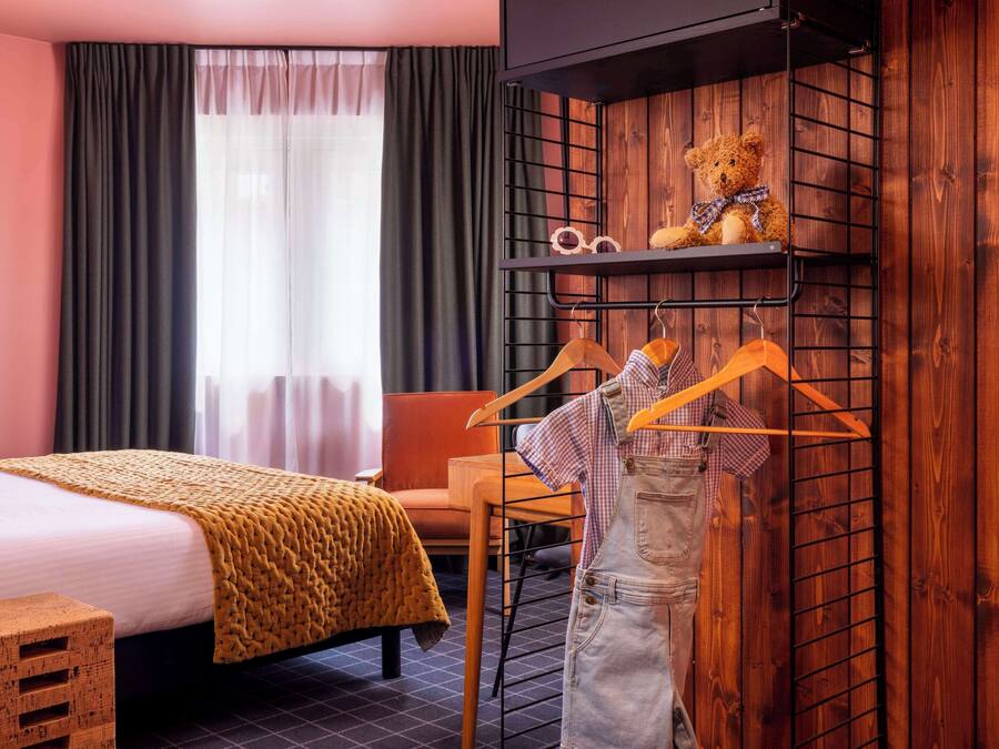 Una habitación de hotel con una cama, una estantería de madera con un oso de peluche y un perchero con ganchos.