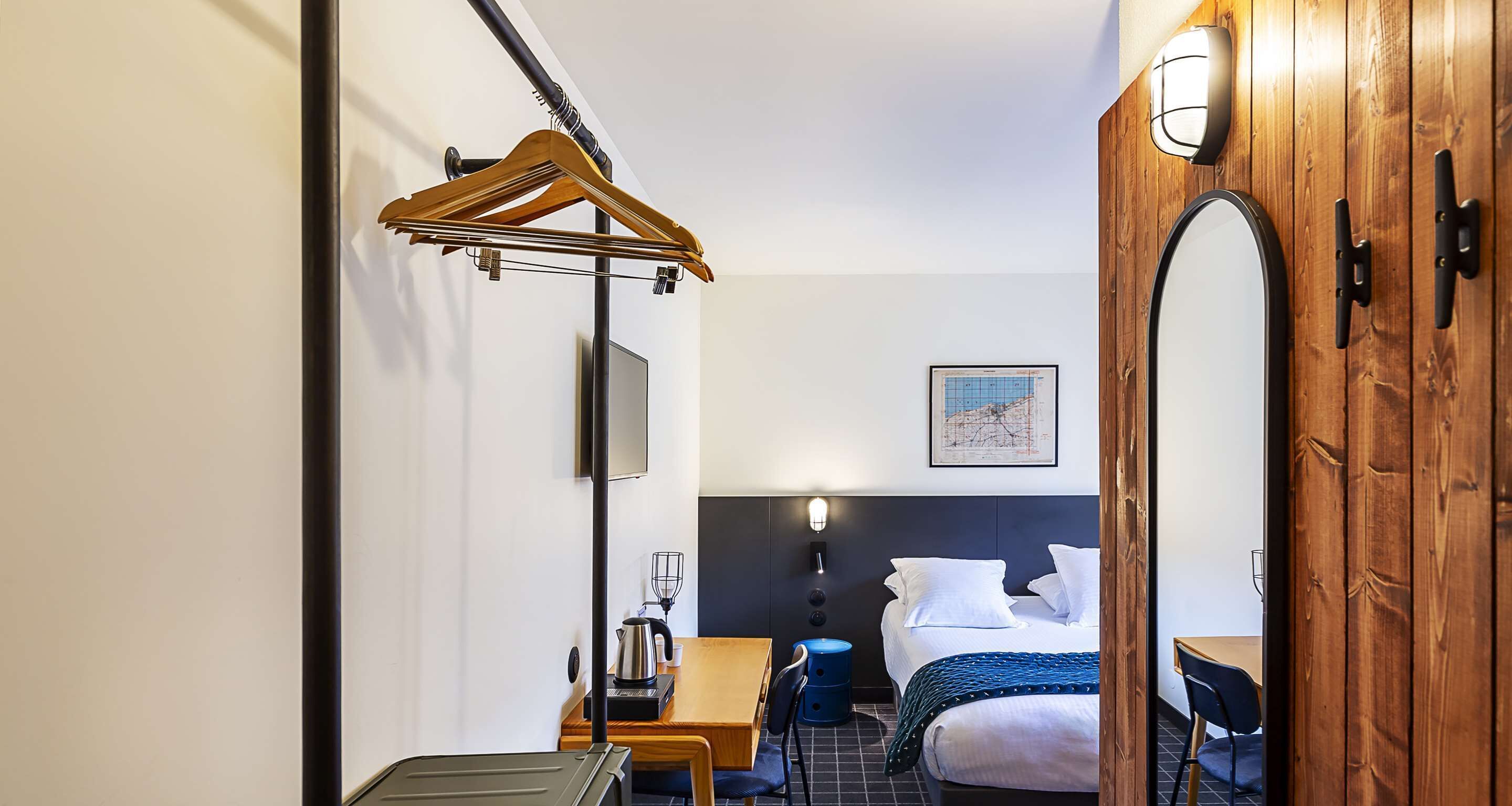 A hotel room with a bed, a wooden dresser, a desk with a chair, a hanging coat rack, and a map on the wall.