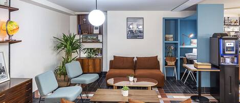 A modern living room with a brown sofa, blue armchairs, and a coffee table. There is a bookshelf with various items and a small desk with a computer.