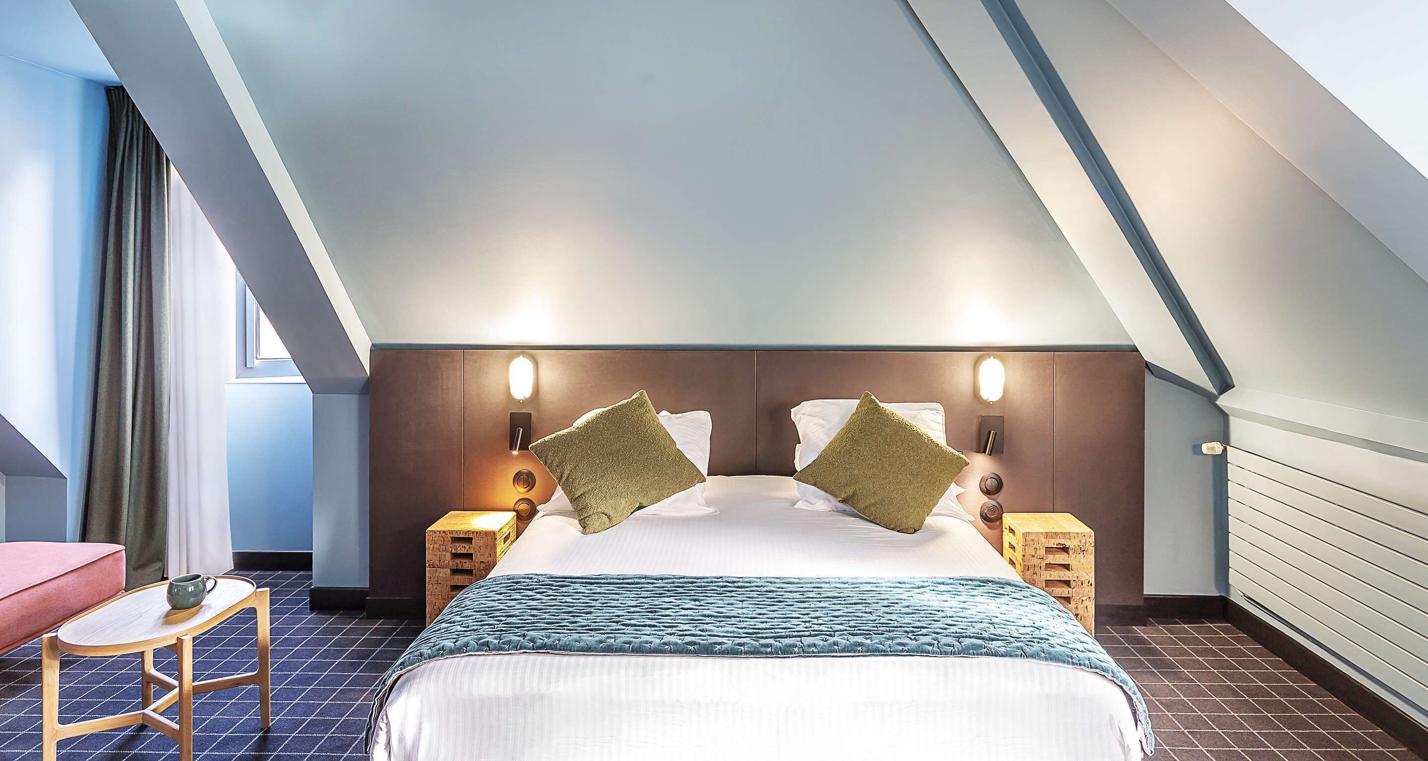 Une chambre d’hôtel avec un plafond en pente, un lit avec deux oreillers, une table de chevet avec une lampe et un petit tabouret en bois.