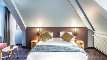 Une chambre d’hôtel avec un plafond en pente, un lit avec deux oreillers, une table de chevet avec une lampe et un petit tabouret en bois.