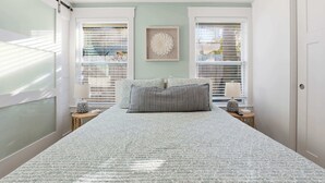A bedroom with a large bed, two bedside tables with lamps, a wall-mounted artwork, and a window with blinds. - Heated Pool + Putting Green | Walk to Beach (Tybee Island)