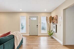 A spacious hallway with a wooden floor, a green sofa with a beige throw, and a door with a decorative glass panel.