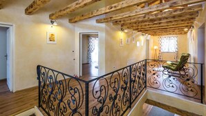 Un interior espacioso con vigas de madera a la vista, una escalera con barandillas negras ornamentadas y una silla verde junto a una ventana.