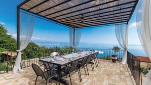 Un área de comedor al aire libre con una mesa dispuesta para seis personas, con vistas a un paisaje marino y una ciudad lejana.