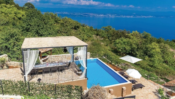 Una piscina en la azotea con vistas al mar, rodeada de exuberante vegetación y un área de descanso cubierta.