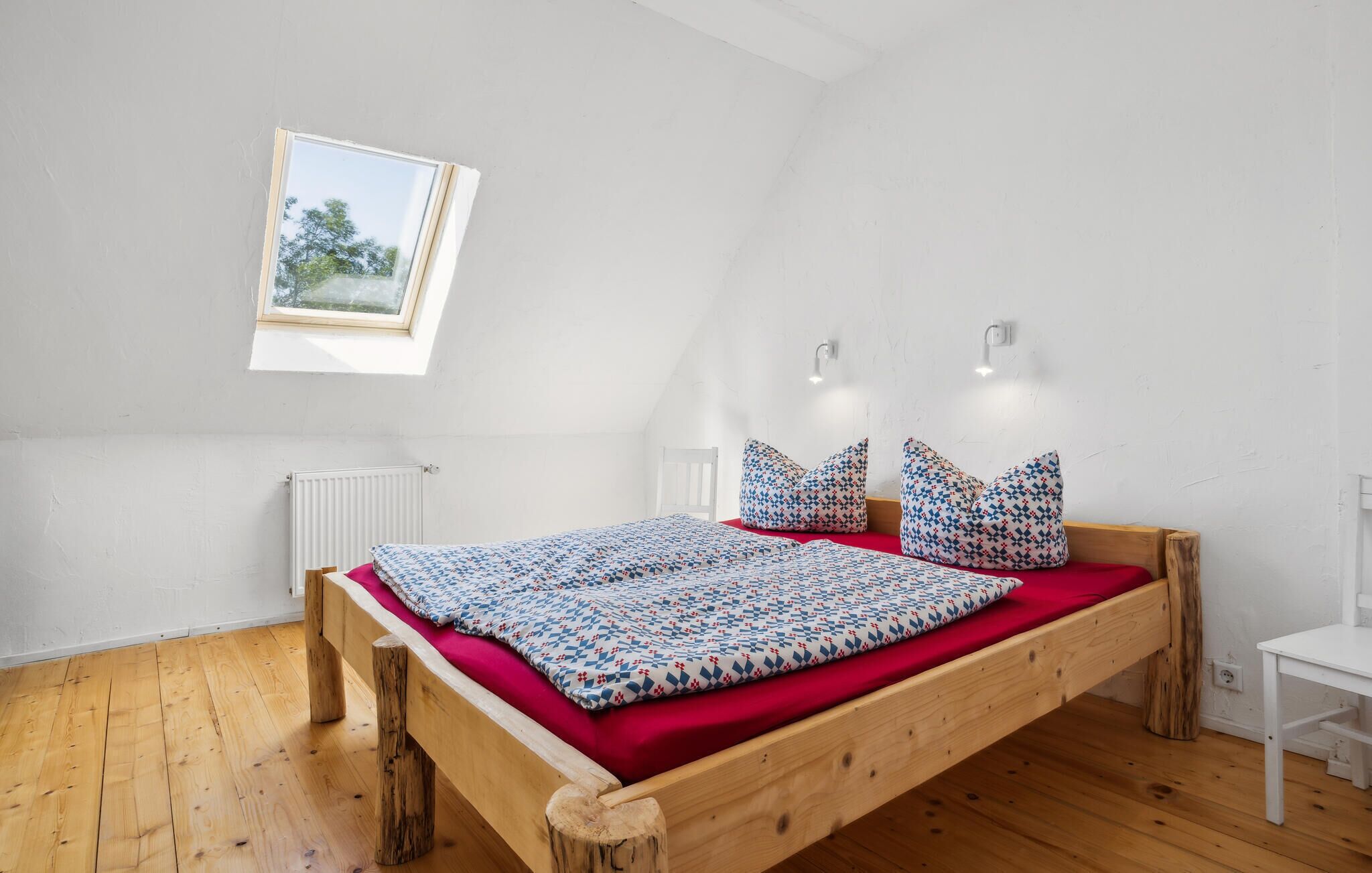 Une chambre avec un plafond en pente, un cadre de lit en bois, un matelas rouge, des coussins à motifs, une petite fenêtre et un radiateur blanc.
