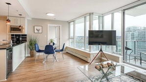 A modern living room with a large window, a flat-screen TV, a dining area with blue chairs, and a kitchen with a microwave.