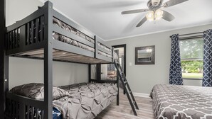 Habitación con literas, ventilador de techo, ventana con cortinas estampadas y puerta que da a otra habitación.