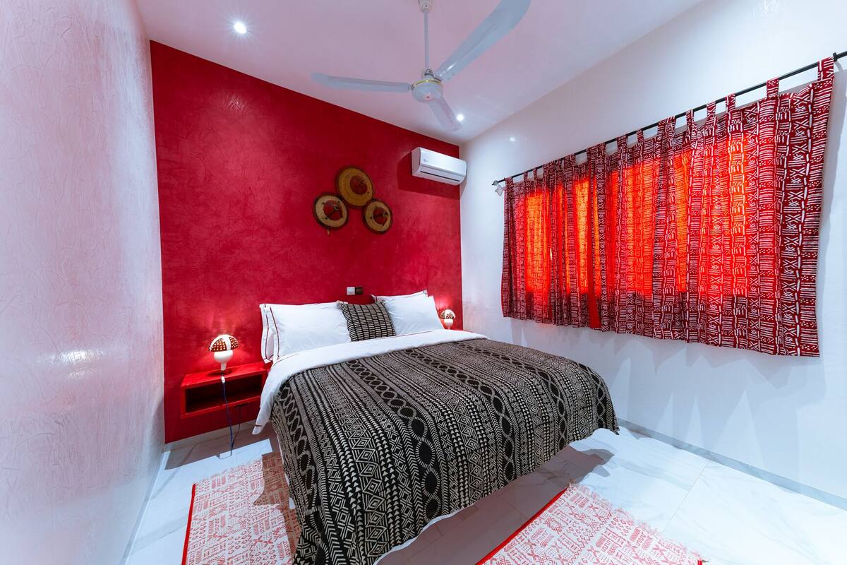 A bedroom with a bed, a ceiling fan, a red wall, and a window with patterned curtains.