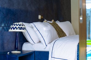 Una cama bien hecha con ropa de cama blanca y detalles azules, una mesita de noche azul con una lámpara, y una pared azul con textura.