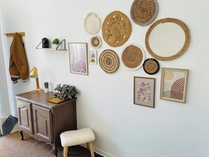 A wall with various woven mats, a framed picture, a small potted plant, a lamp, and a bench.