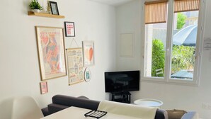 Un salon moderne avec une table blanche, deux chaises blanches, un canapé noir, un téléviseur et des œuvres d’art accrochées au mur.