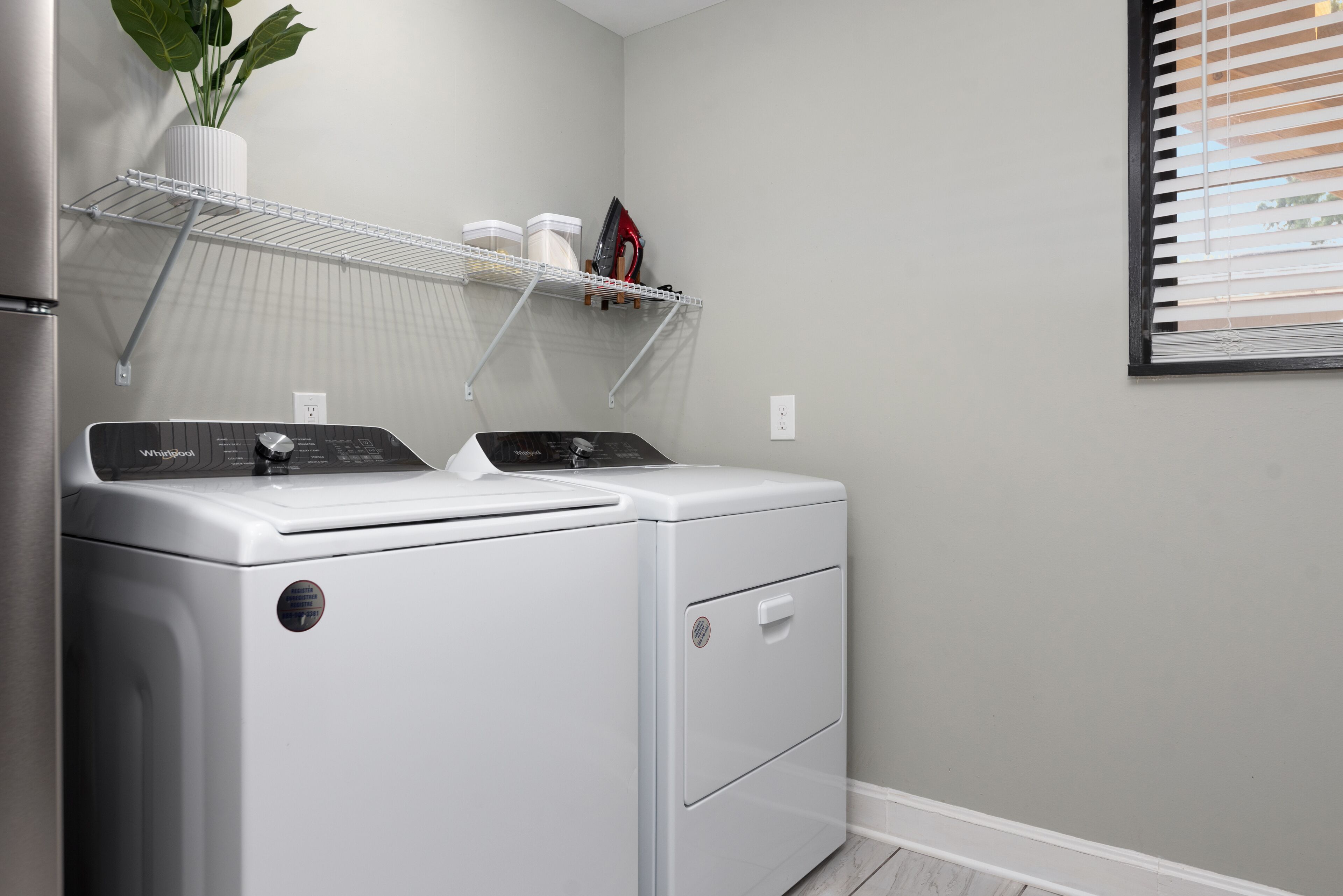 A laundry area with two stacked washers and dryers, a white shelf with containers, and a potted plant.