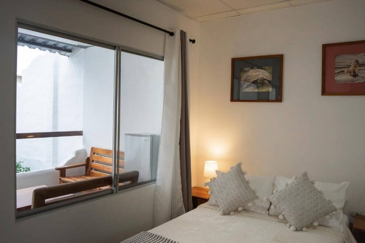 Een hotelkamer met een bed, een nachtkastje, een lamp, een raam met uitzicht op een balkon en twee prestatiekaders aan de muur.