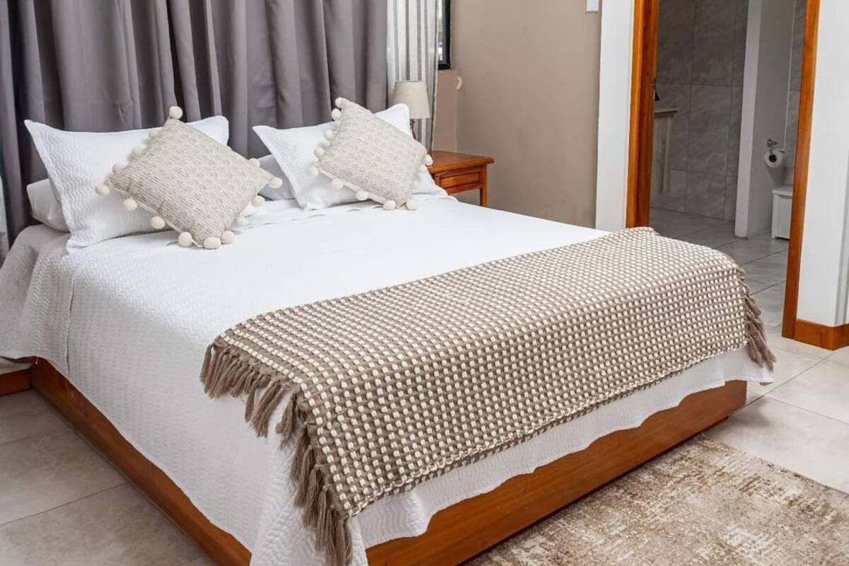 Een moderne slaapkamer met een groot bed, grijze gordijnen, een houten nachtkastje en een badkamer door een open deur zichtbaar.