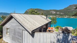Una cabaña de madera con techo inclinado, un pequeño porche con mesa y sillas, y vista a un lago azul con montañas al fondo.