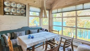Una habitación acogedora con techo de madera, una mesa de comedor con mantel blanco, cojines estampados en azul y blanco, y vistas a una extensión de agua azul a través de amplias ventanas.