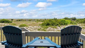 Een terras met twee Adirondack-stoelen en een tafel, met uitzicht op een natuurlandschap met bomen en duinen.