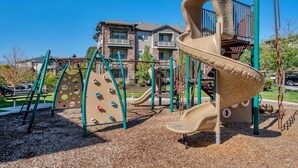 Ein Spielplatz mit Klettergerüsten und Rutschen, umgeben von einem Kiesbereich und Wohnhäusern im Hintergrund.