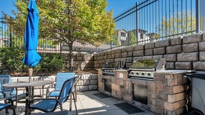 Eine Terrasse im Freien mit einem Stein-Barbecue, blauen Stühlen und einem Tisch mit blauem Sonnenschirm.