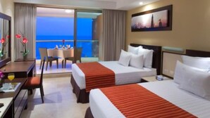 A hotel room with two beds, a desk, a chair, and a balcony with a view of the sea.