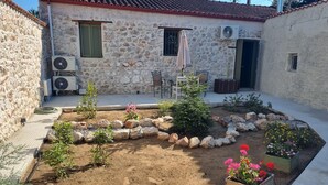 Une maison en pierre avec un toit en tuiles, un espace extérieur aménagé pour s’asseoir et un jardin où poussent différentes plantes et fleurs.