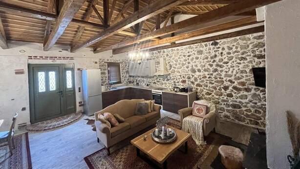 Un intérieur rustique avec des poutres apparentes, un mur en pierre, un coin cuisine et un espace salon confortable avec un canapé et des fauteuils.