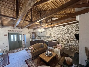 A rustic interior with exposed wooden beams, a stone wall, a kitchenette, and a cozy seating area with a sofa and armchairs. - We Created a Completely Functional Quest House Based on Stone and Wood, (Aliartos)