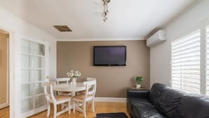 A living room with a black leather sofa, a white dining table with chairs, a flat-screen TV mounted on the wall, and a sliding glass door leading outside. - Bali in the Barossa (Gawler West)