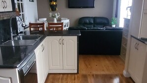 A modern kitchen with white cabinetry, a black countertop, and a wooden floor. There is a dining area with a black sofa and a small table. - Bali in the Barossa (Gawler West)