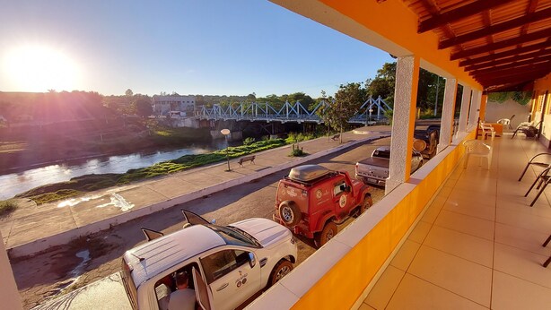 A view from a balcony overlooking a river, a bridge, and a parking area with a white van and a red vehicle. - POUSADA BEIRA RIO - JALAPÃO (Ponte Alta do Tocantins)