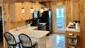 A wooden kitchen with a central island, two bar stools, a refrigerator, a stove, and hanging pendant lights.