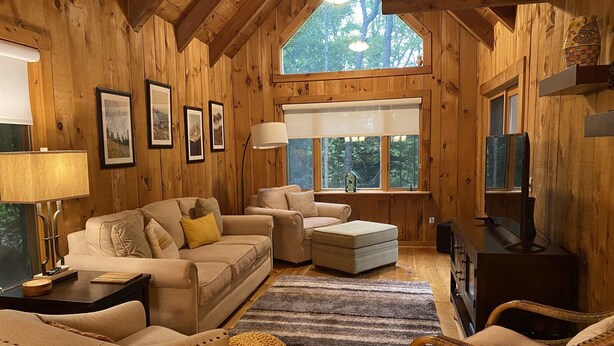A cozy wooden cabin interior with a sofa, ottoman, wicker chair, and a television.