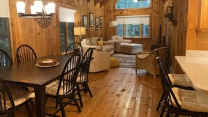 A cozy cabin interior with a dining area, a living room with a sofa and ottoman, and a large window providing a view of the outdoors.