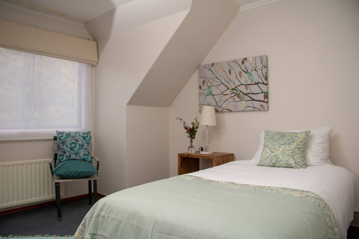 Ein Schlafzimmer mit Bett, Stuhl, Nachttisch, Lampe und einem Fenster mit Jalousien.