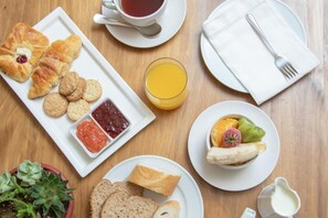 A breakfast setting with croissants, biscuits, jam, a glass of orange juice, a fruit bowl, and a small potted plant.