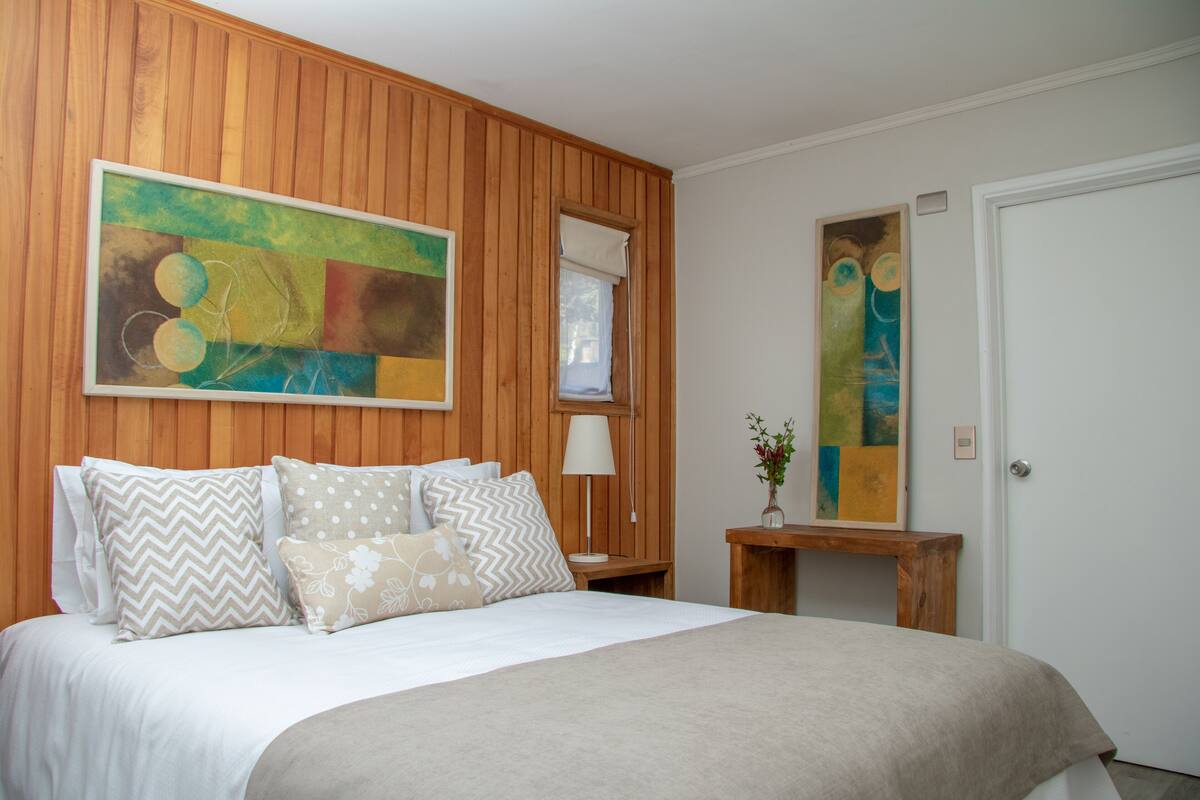 Ein Schlafzimmer mit einer holzverkleideten Wand, einem Bett mit vielen Kissen, einem Nachttisch mit Lampe und zwei abstrakten Bildern.