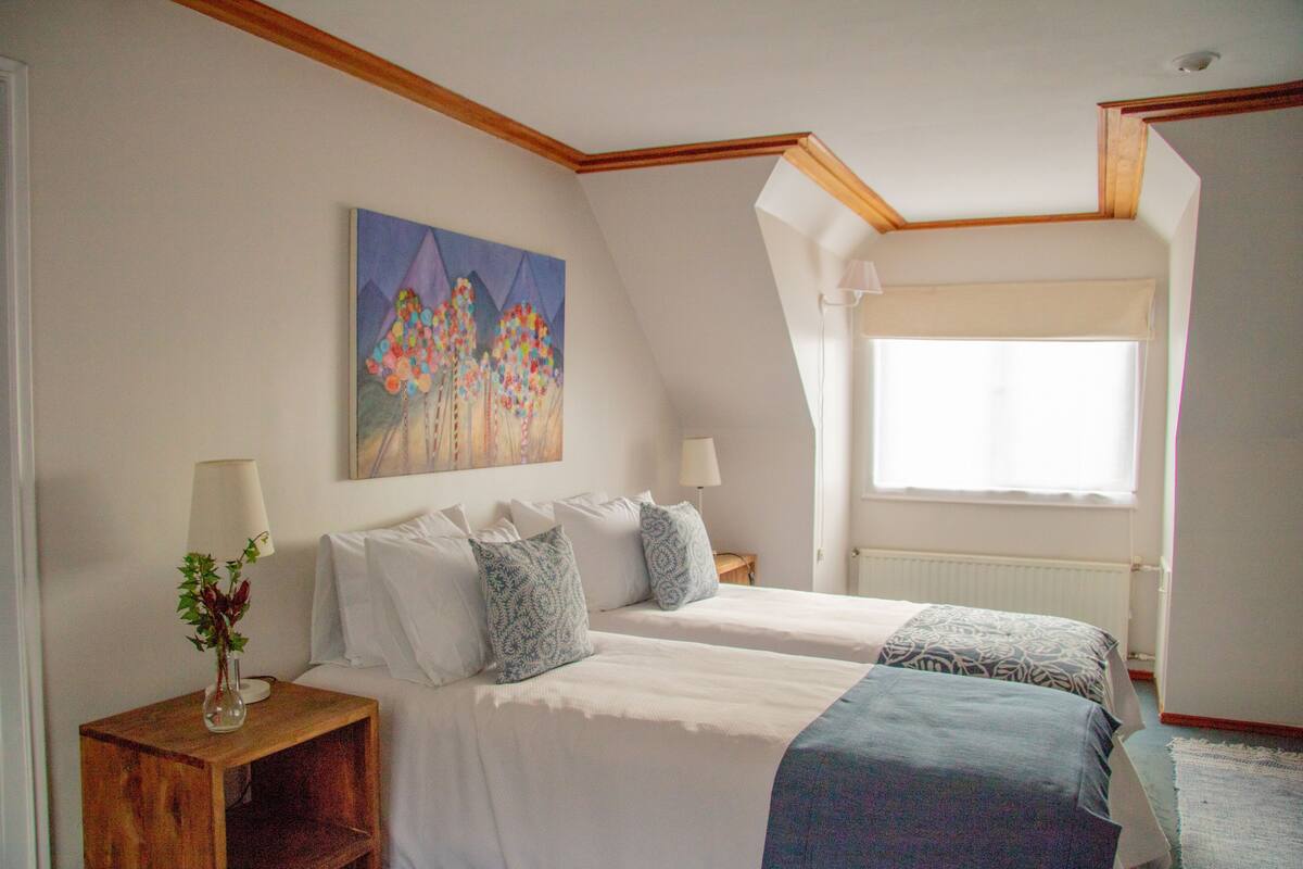 Ein Schlafzimmer mit zwei Betten, einem Bild an der Wand, einer Lampe auf einem Nachttisch und einem Fenster mit Aussicht.