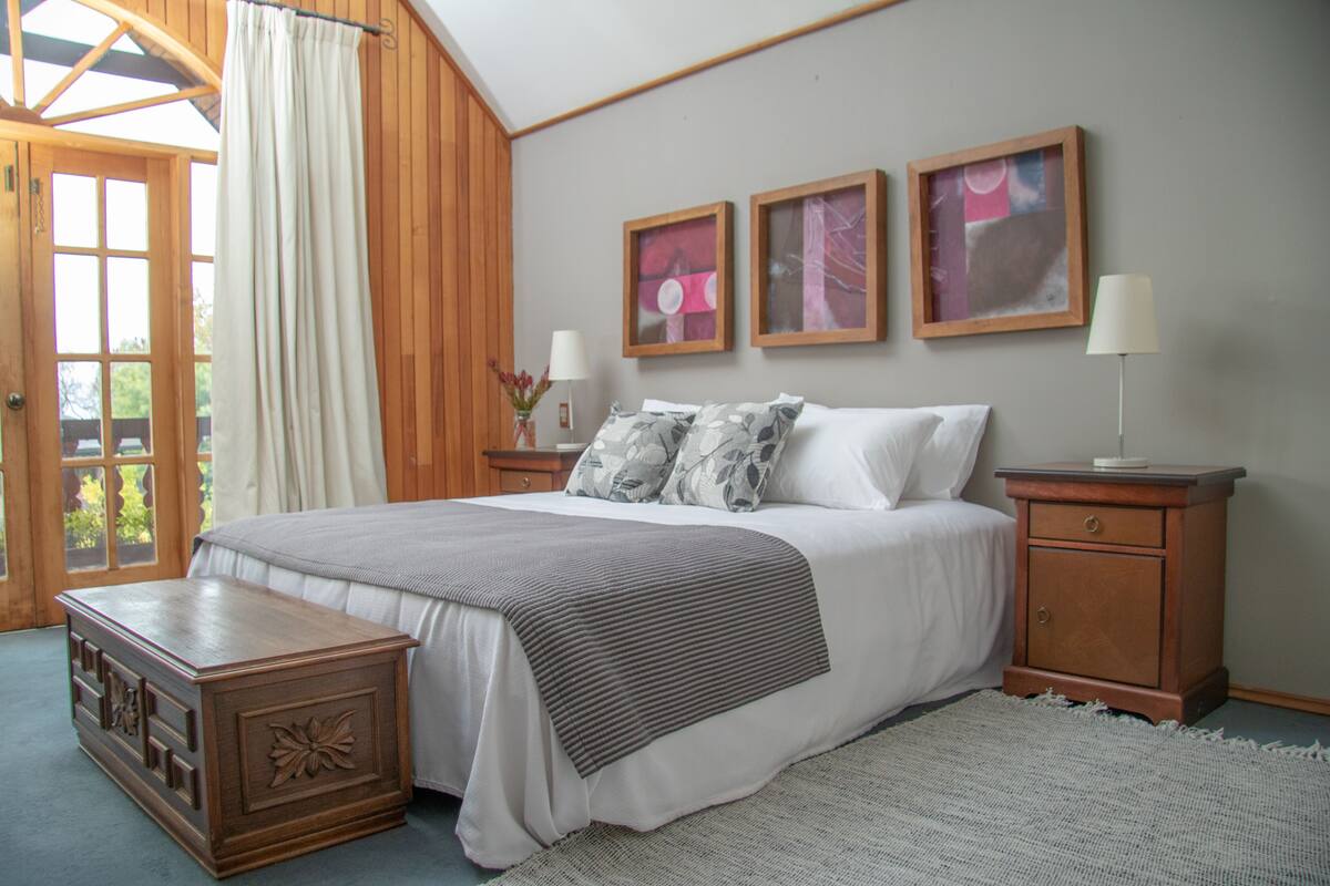 Ein Schlafzimmer mit einem großen Bett, Nachttischen aus Holz, einer Kommode und Blick durch ein Fenster mit Vorhängen.