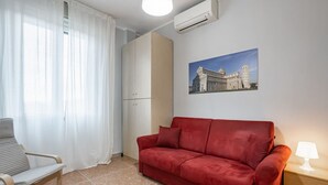 Ein Zimmer mit einem roten Sofa, einem Holzstuhl, einem Nachttisch mit Lampe, einem Bild eines Gebäudes und einer Klimaanlage.