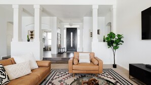 A spacious living room with a brown leather sofa, a matching armchair, and a glass coffee table. A large plant and a black entertainment unit are visible. - Gorgeous Lakefront Home with Infinity Pool & Hot Tub, Sleeps Up to 12 (Glendale)