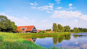 Een traditioneel Nederlands huis met een rood dak, gelegen naast een rustig meer met weelderige begroeiing en bomen op de achtergrond.