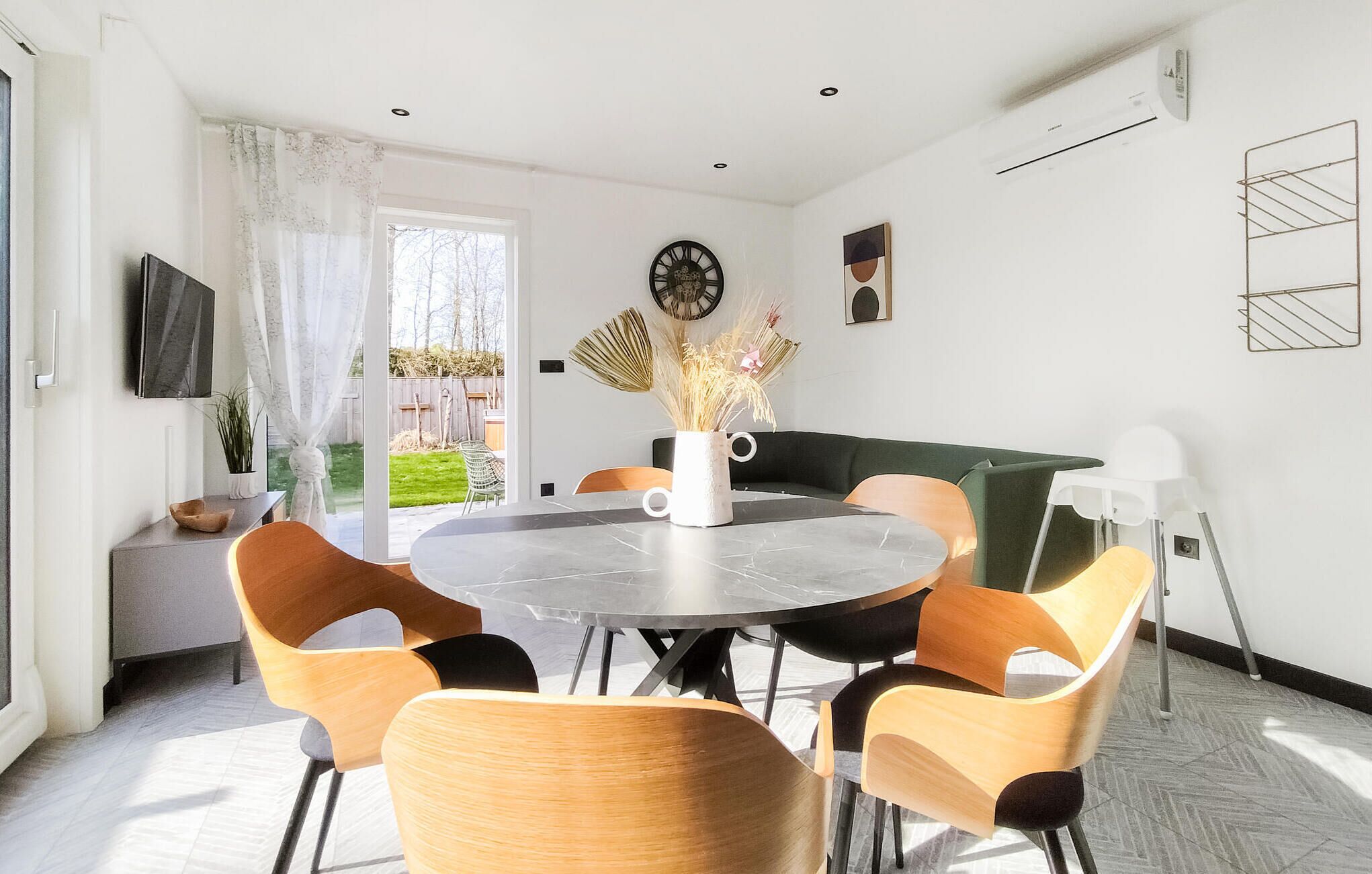 Un espace repas moderne avec une table ronde, six chaises, un vase contenant des plantes séchées, une horloge et un canapé vert.
