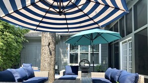 A covered outdoor seating area with wicker furniture and blue cushions, a large striped umbrella, and a decorative elephant figure on a table.