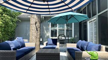 Un área de estar al aire libre techada con muebles de mimbre y cojines azules, un paraguas ancho a rayas y una figura decorativa de elefante sobre una mesa.