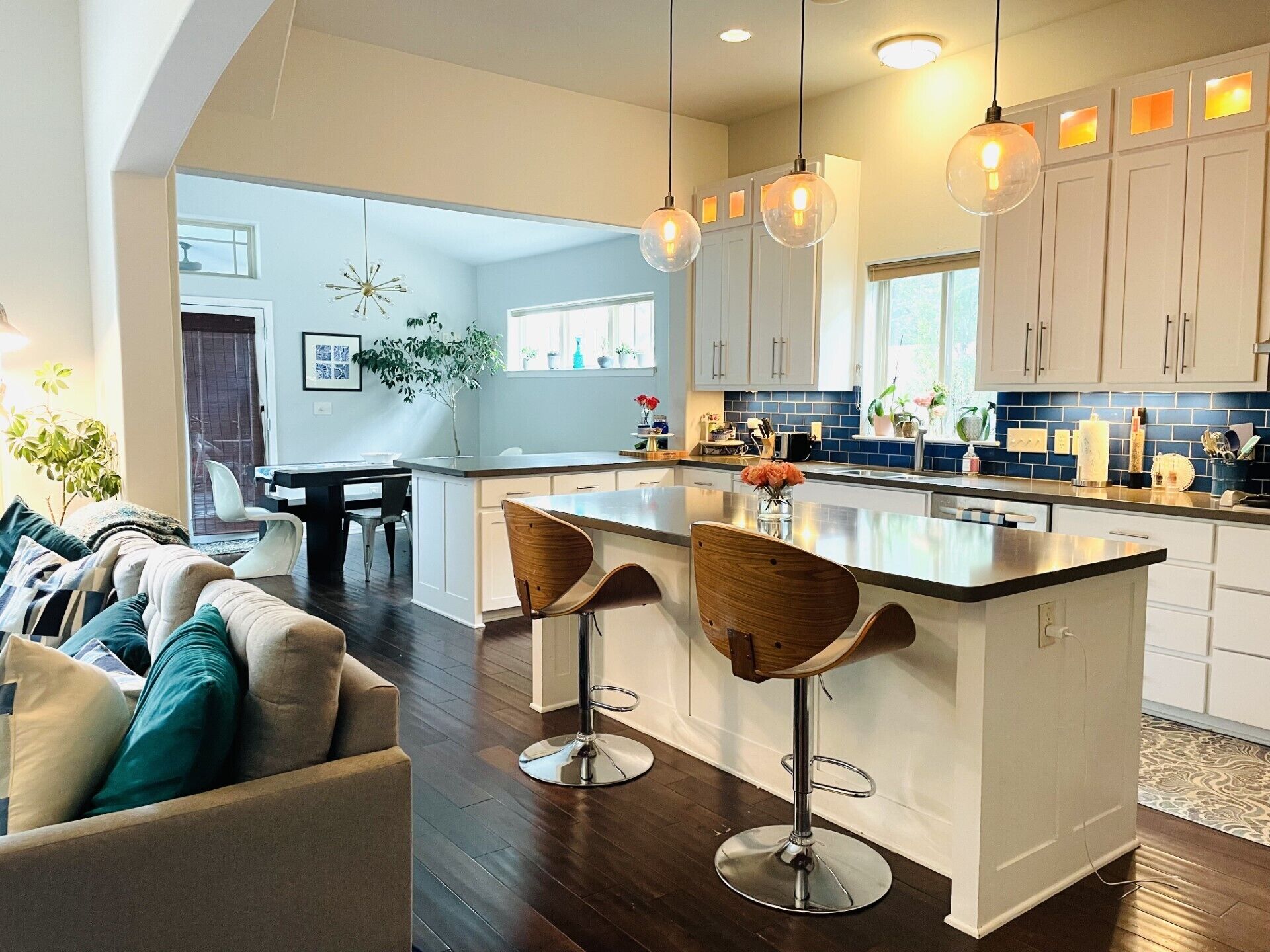 Una cocina moderna con armarios blancos, una isla central con taburetes, iluminación colgante y una zona de comedor con sofá y sillas.
