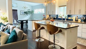 A modern kitchen with white cabinets, a central island with stools, pendant lighting, and a dining area with a sofa and chairs.